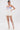 A woman poses in a blue and white strapless corset top with pearl buttons, the GUESS WHO MINI SKORT featuring sculptural pleats, high-waisted shorts, knee-high strappy white heeled sandals, and a pearl choker necklace.