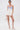 A woman poses against a white background wearing the GUESS WHO MINI SKORT, a high-rise white miniskirt, paired with a strapless blue and white top, knee-high gladiator boots, pearl necklace, and holding a white handbag.
