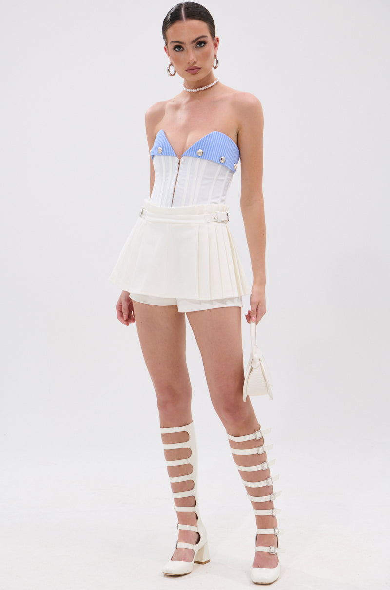 A woman wears the GUESS WHO MINI SKORT—a white pleated mini skort with sculptural details—paired with a blue and white corset top, knee-high strappy boots, pearl necklace, and a small white purse against a white background.
