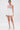 A woman wears the GUESS WHO MINI SKORT—a white pleated mini skort with sculptural details—paired with a blue and white corset top, knee-high strappy boots, pearl necklace, and a small white purse against a white background.