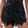 Someone is wearing the GOING OUT LACE TRIM LEATHER SHORTS, high-waisted faux leather shorts with lace trim at the hem and pocket flaps, styled with a sheer black lace top.