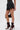A person wears GOING OUT LACE TRIM LEATHER SHORTS, a lacy black top, lace-up high-heeled boots, and holds a small black purse with gold charms. The chic look is photographed from mid-torso to feet.
