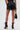 Wearing the GOING OUT LACE TRIM LEATHER SHORTS, paired with a black handbag and maroon heeled lace-up boots, this rear-view image shows the standout faux leather shorts against a plain light background.
