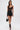 A woman in a sheer black lace top, GOING OUT LACE TRIM LEATHER SHORTS, lace-up high heel sandals, gold hoop earrings, a gold arm cuff, and carrying a black handbag walks forward against a white background.