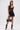 A woman poses against a plain white background wearing high-waisted GOING OUT LACE TRIM LEATHER SHORTS, a black lace bodysuit, lace-up heeled boots, large hoop earrings, and holding a black handbag with gold accents.