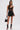 A woman stands against a plain background wearing GOING OUT LACE TRIM LEATHER SHORTS, a black lace bodysuit, lace-up peep-toe boots, large hoop earrings, and holding a black handbag.