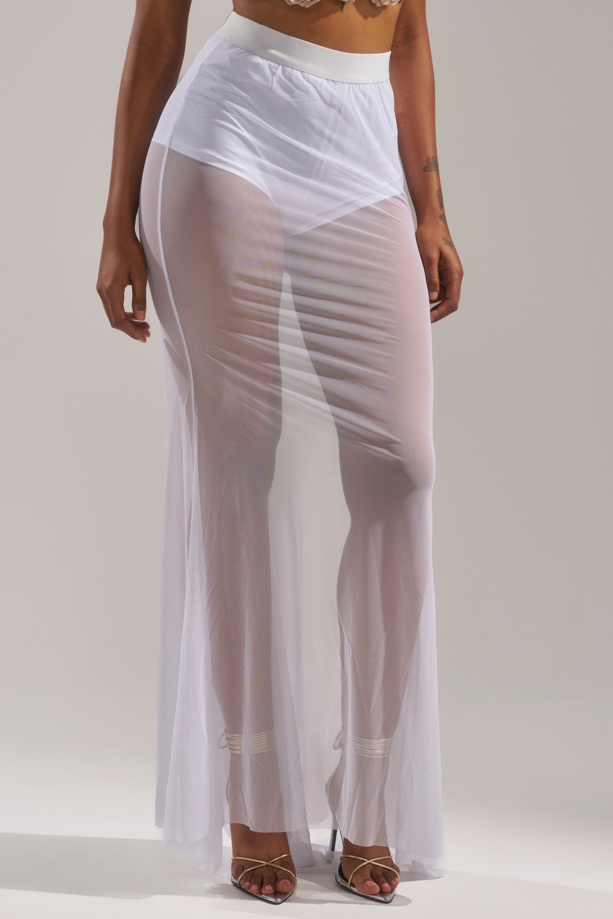 A person wears the DANCING QUEEN MESH MAXI SKIRT IN WHITE, a sheer maxi with a high-rise elastic waistband and visible white underwear, styled with heeled sandals for an effortlessly chic festival look against a neutral background. 1