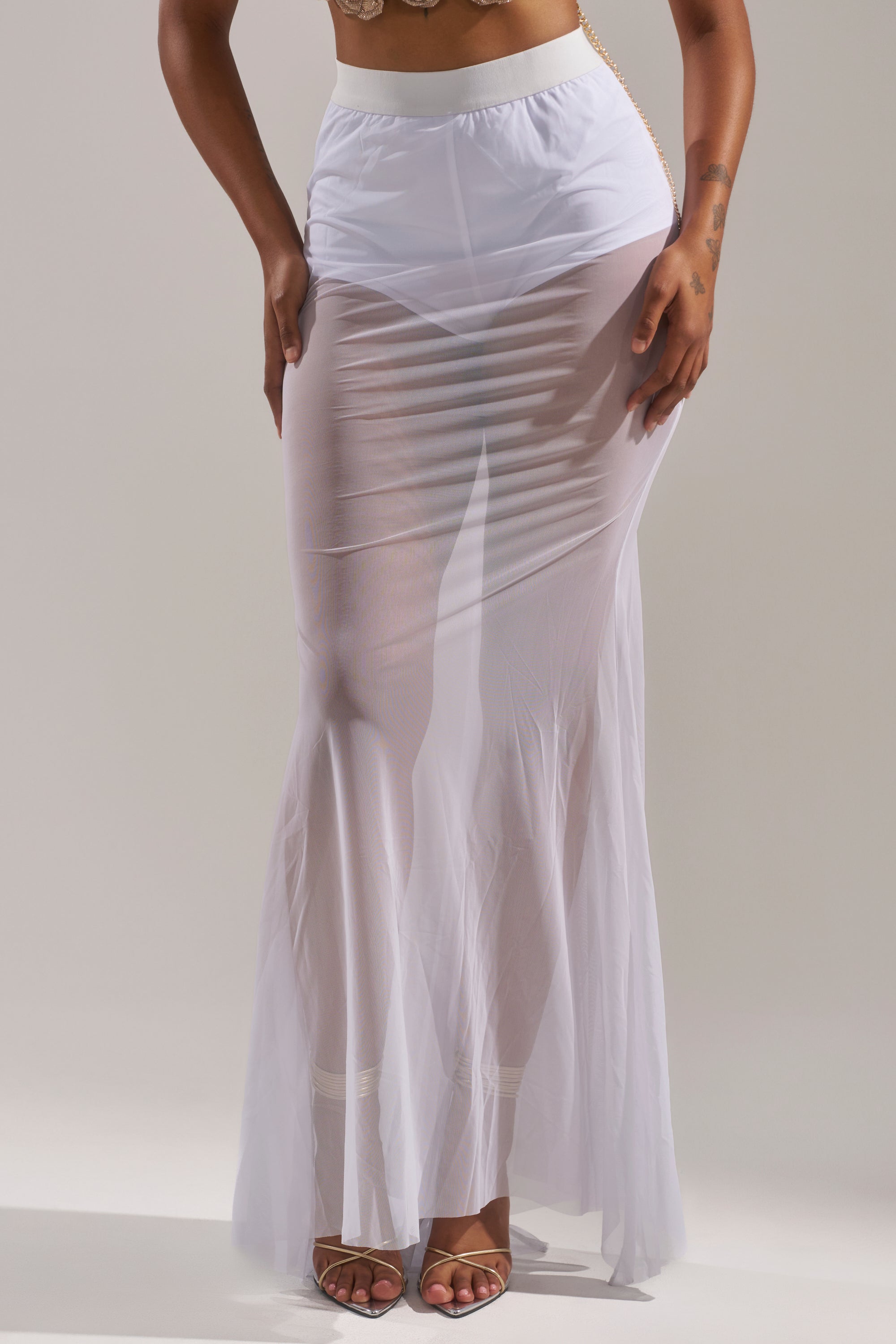 A person models the DANCING QUEEN MESH MAXI SKIRT IN WHITE—a long, sheer mesh maxi skirt with a high-rise elastic waistband—standing with one hand on their hip and the other lifting part of the skirt, paired with strappy high-heeled sandals. 2
