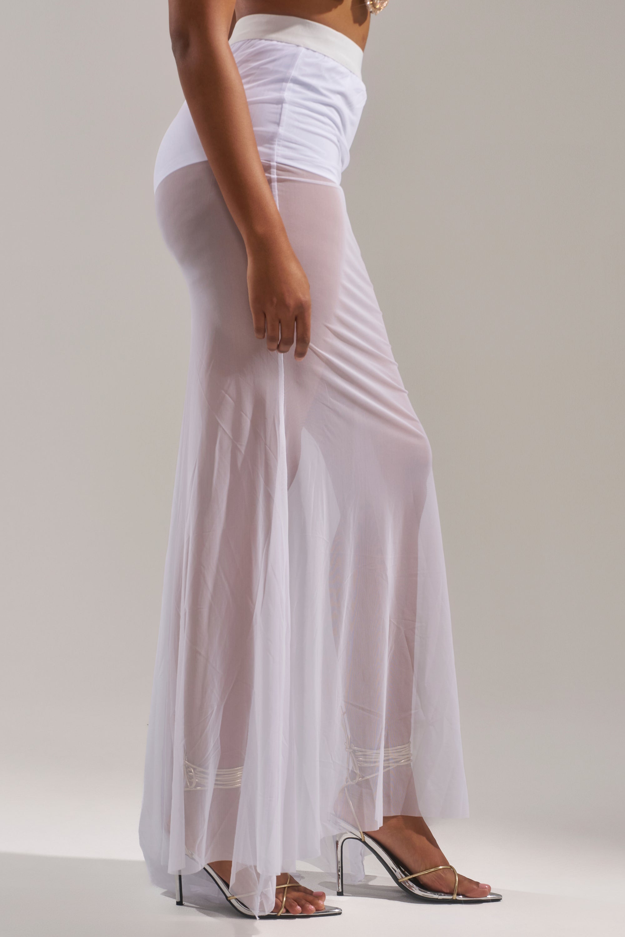 A person wears the DANCING QUEEN MESH MAXI SKIRT IN WHITE, featuring a sheer mesh fabric and high-rise elastic waistband, styled with silver high-heeled sandals against a plain background—ideal festival outfit inspiration. 3