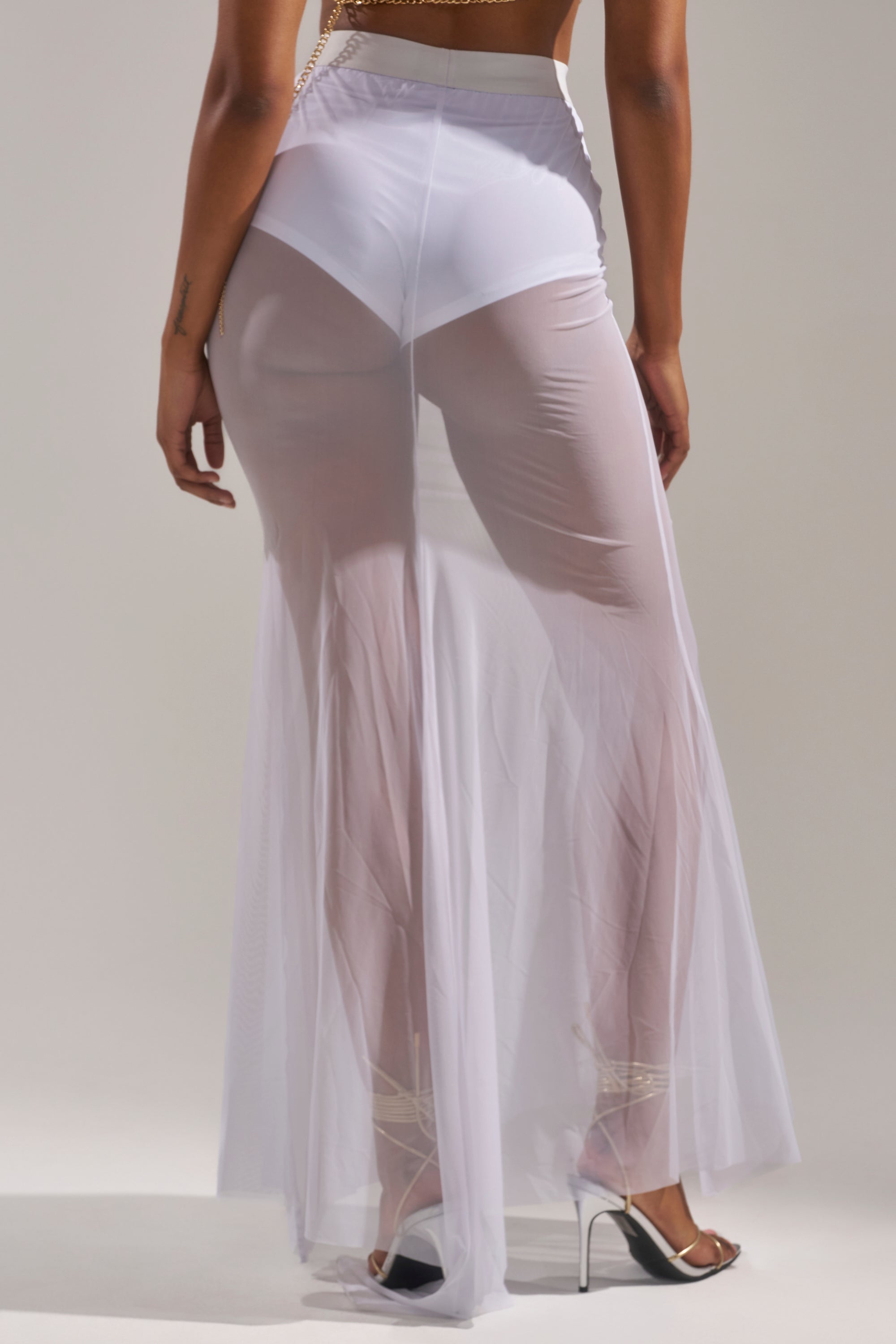 A person faces away, wearing the DANCING QUEEN MESH MAXI SKIRT IN WHITE—a sheer, high-rise maxi skirt over white underwear and heels. The airy mesh flows from waist to floor, adding a dreamy touch ideal for festival styles. 4