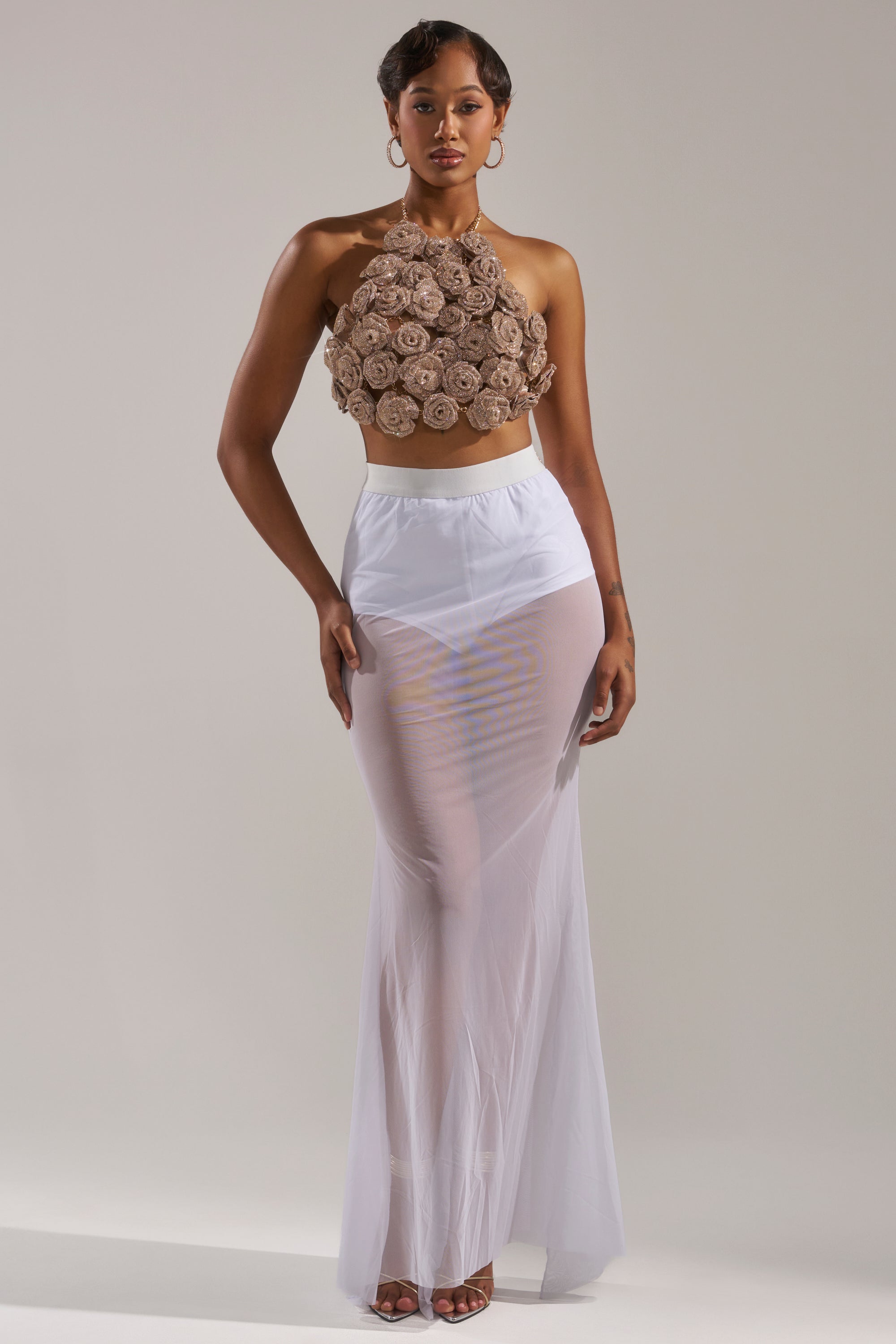 A woman poses against a plain light background, wearing the DANCING QUEEN MESH MAXI SKIRT IN WHITE—a long mesh maxi skirt with a high-rise elastic waistband styled over white undergarments, paired with a sleeveless brown halter crop top. 5