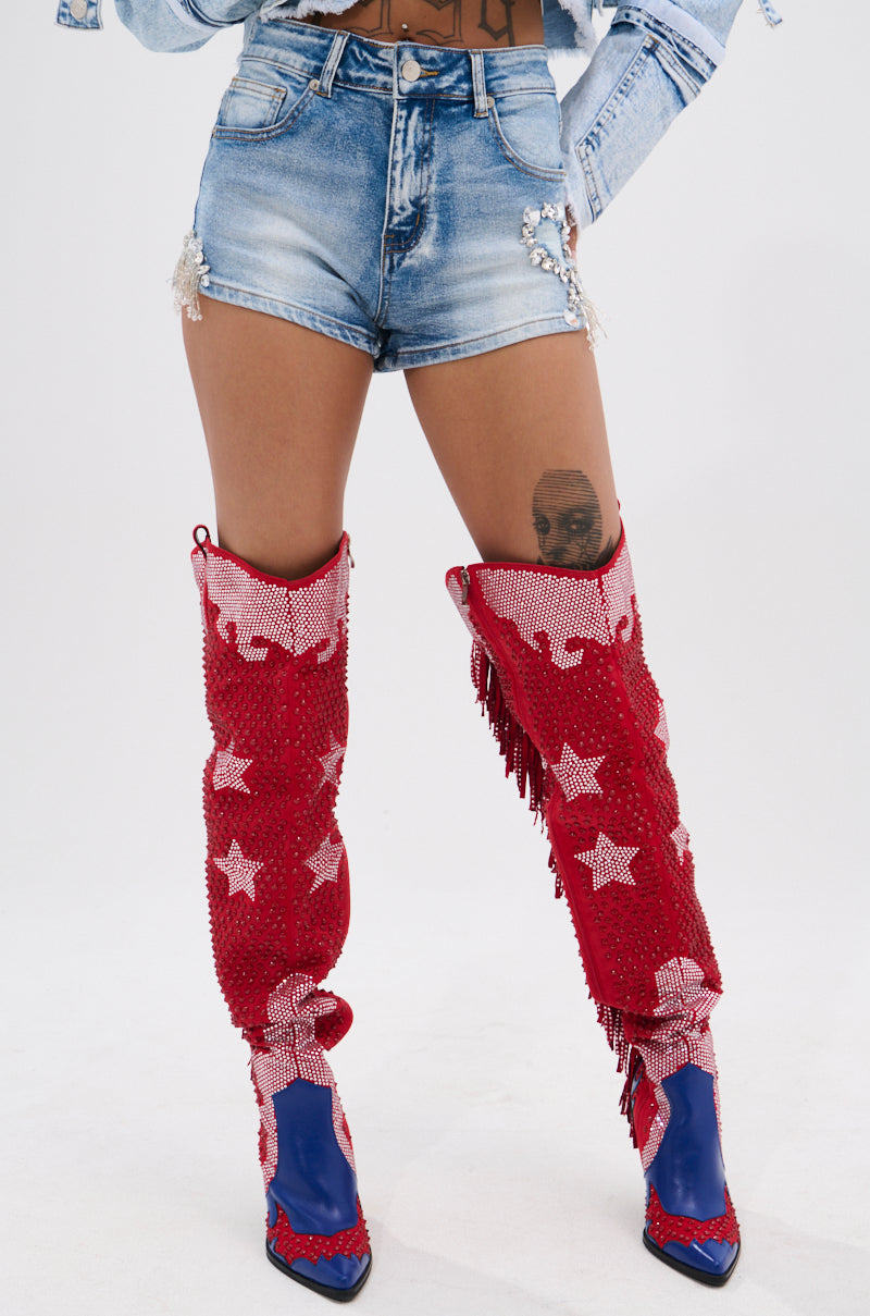 A person wearing the KYLIE EMBELLISHED DENIM SHORT with distressed details and knee-high red boots with white stars and blue pointed toes stands against a plain white background, showing a tattoo on their left thigh. 2