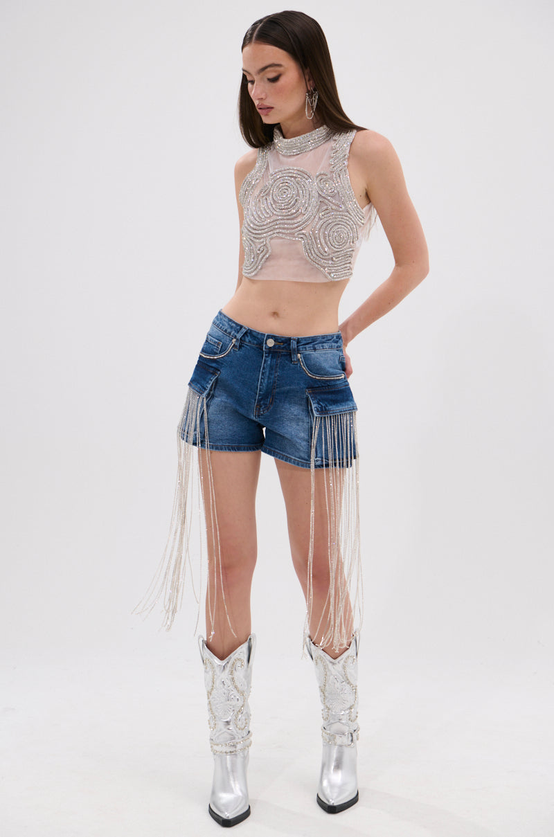 A woman poses against a plain white background, showcasing streetwear style in DANCING ALL NIGHT LONG DENIM EMBELLISHED SHORTS with long rhinestone fringe, paired with a sleeveless beaded crop top and metallic silver cowboy boots. 7