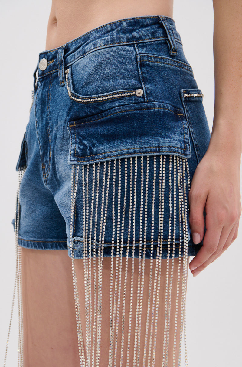 Close-up of a person wearing the DANCING ALL NIGHT LONG DENIM EMBELLISHED SHORT, featuring large front pockets and long rhinestone fringe that extends past the hem for a shimmering, streetwear-inspired look. The person's right arm is at their side. 8
