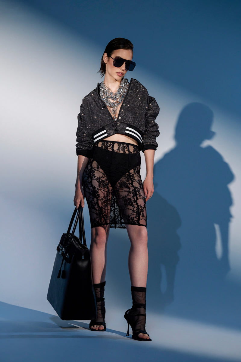 A woman poses against a blue background wearing sunglasses, a sparkly black varsity jacket, a layered necklace, heeled sandals, and holding a large black bag. She completes her look with the BABY PLEASE PLEASE LACE MIDI SKIRT over black underwear. 2