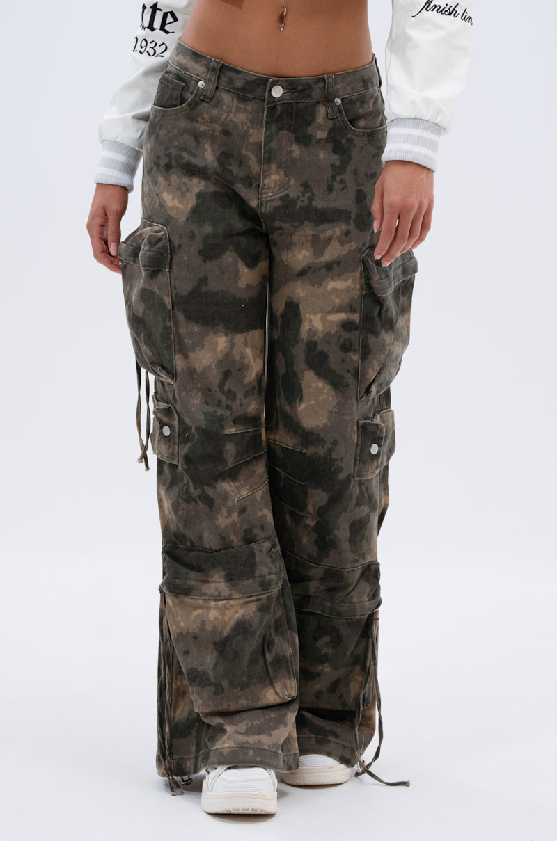 A person wears the DOWN AND DIRTY CARGO PANT—high-rise relaxed fit camo cargos with side pockets, drawstrings, and snap details—paired with white sneakers and a cropped white jacket. 2
