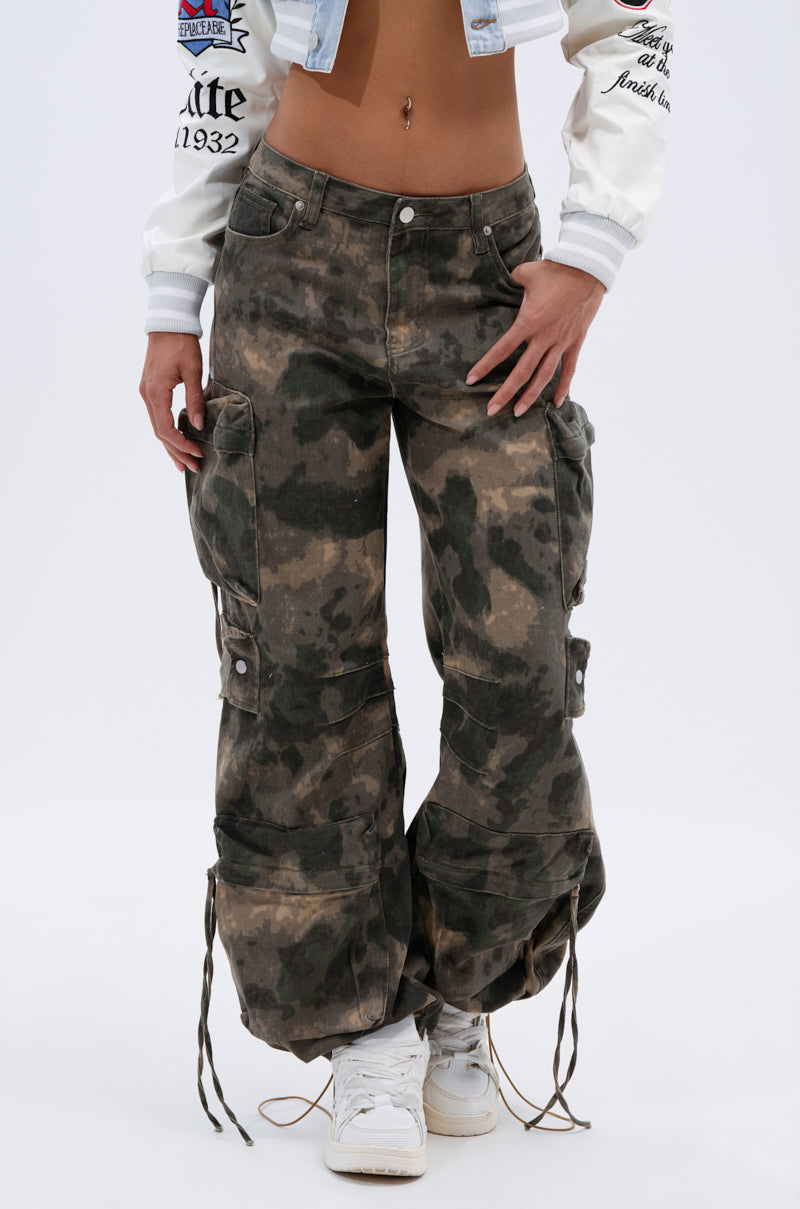 A person wears the DOWN AND DIRTY CARGO PANT—high-rise, relaxed fit, camouflage with drawstring hems—paired with white sneakers and a cropped white jacket revealing their midriff and belly button piercing, set against a plain light background. 3