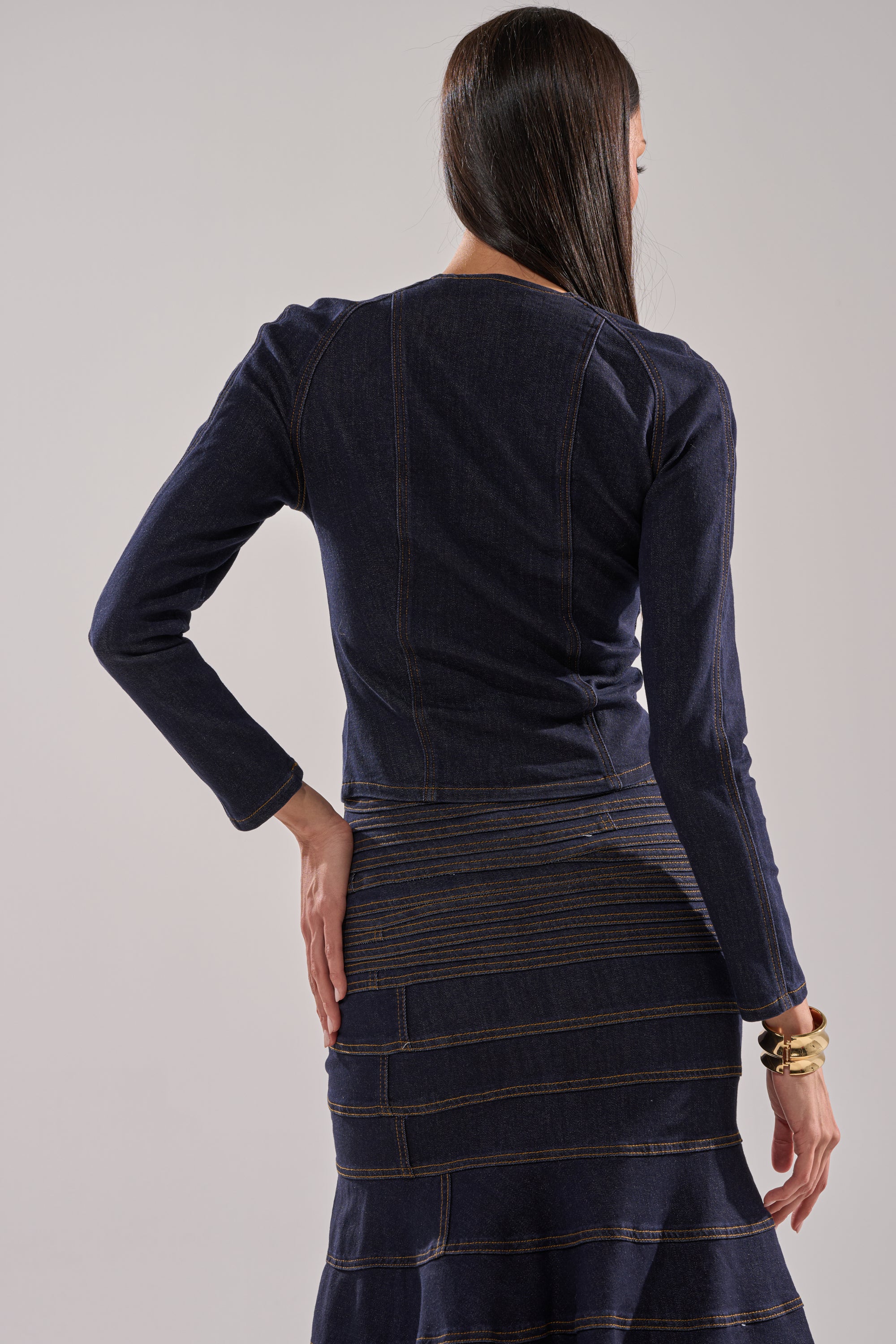A woman with long, straight hair stands facing away, wearing the ALL ABOUT THAT DENIM BLOUSE—a dark-wash, long-sleeve denim top with contrast stitching. One hand on her hip displays gold bracelets on her wrist. 4