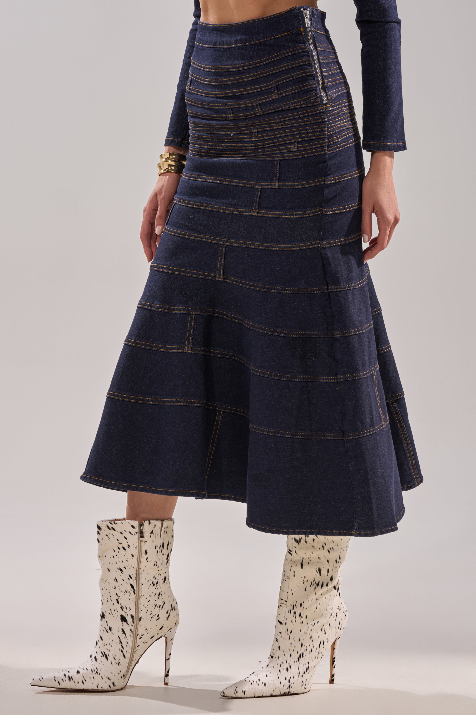 A person wearing the ALL ABOUT THAT DENIM MAXI SKIRT—a navy blue, high-waisted A-line maxi with visible stitching—paired with a matching long-sleeve top and white knee-high boots featuring black speckles and pointed toes. 3