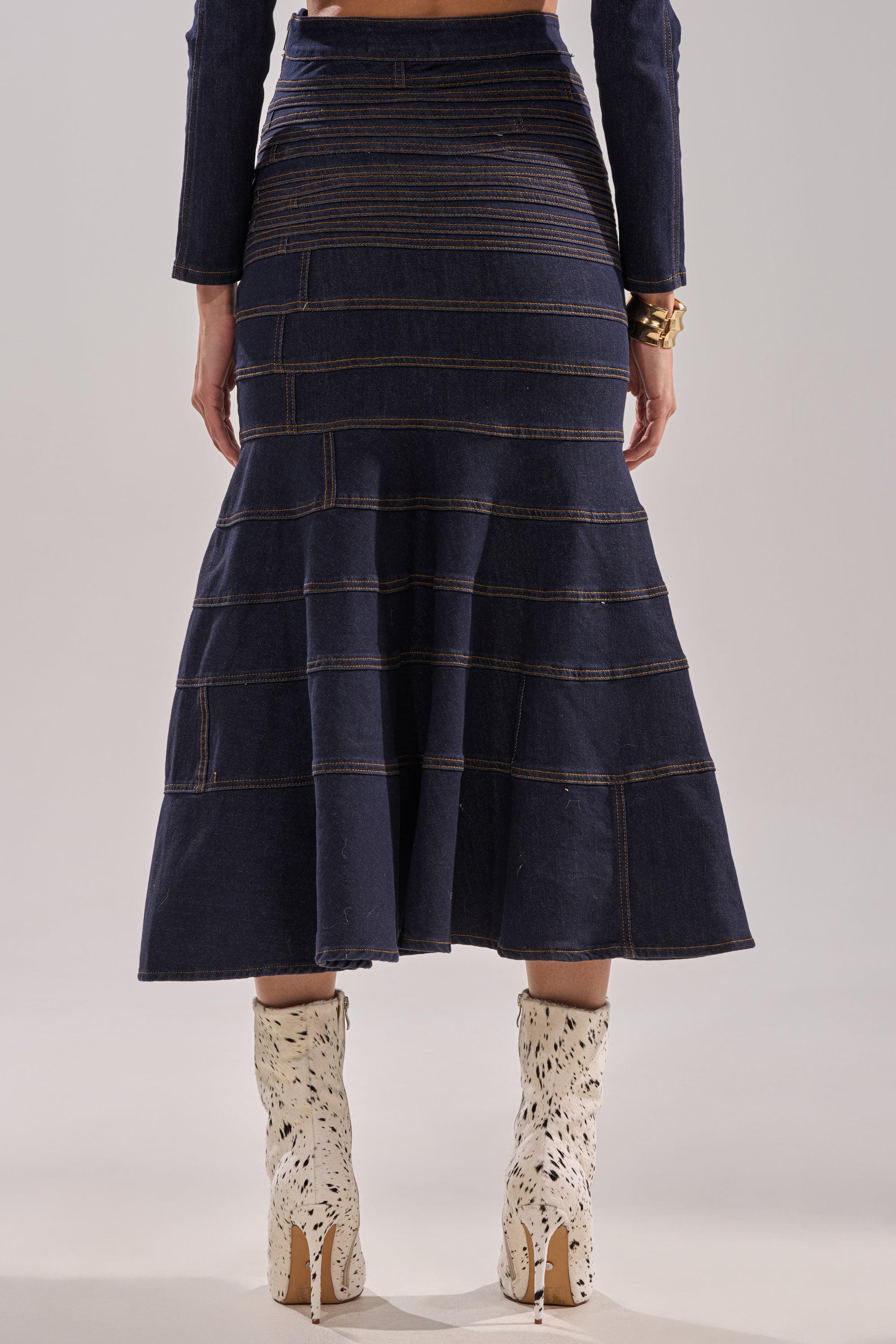 A person is seen from the back wearing the ALL ABOUT THAT DENIM MAXI SKIRT, a dark-wash, high-waisted denim skirt with horizontal seams, paired with light-patterned mid-calf boots against a neutral backdrop. 4