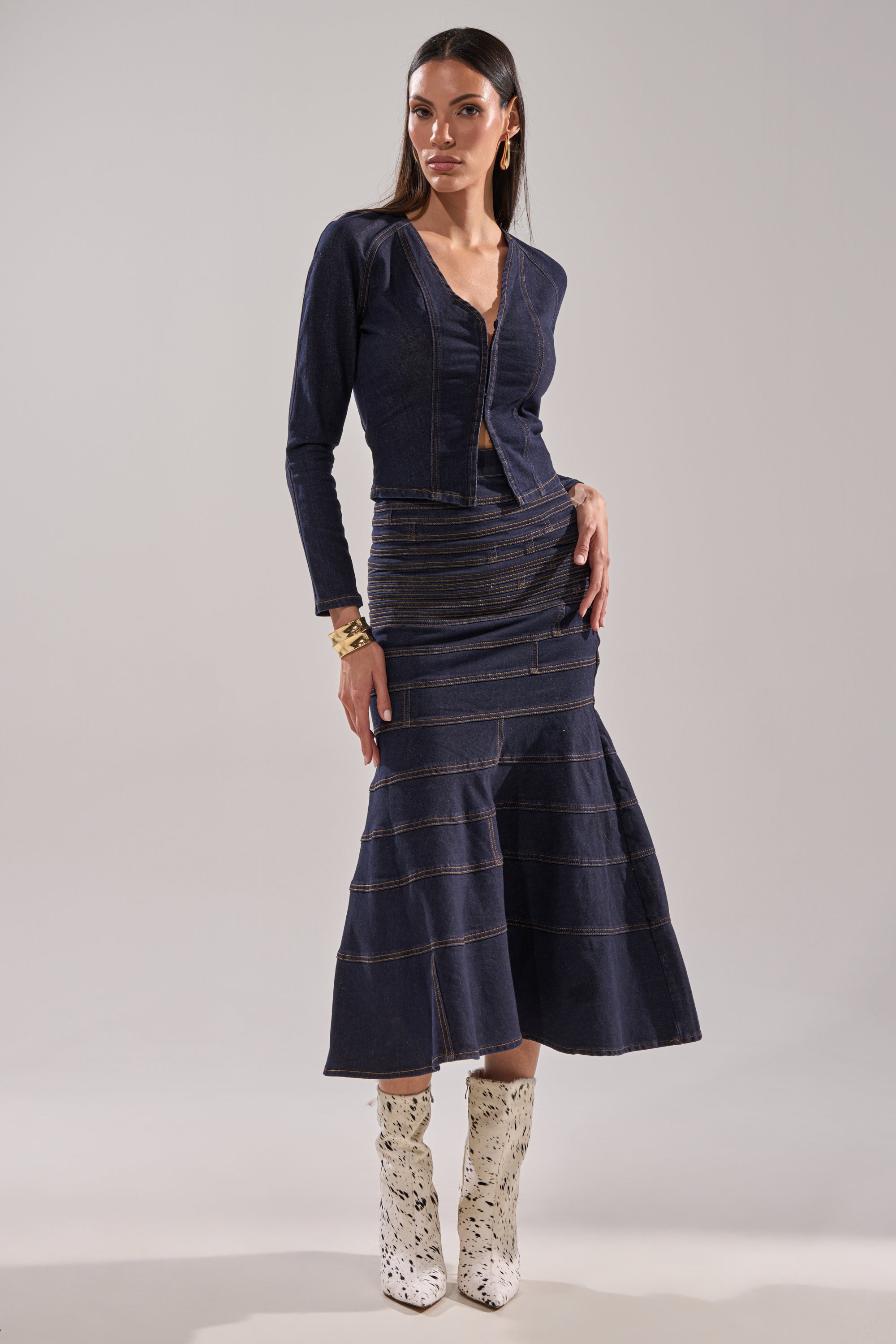 A woman poses against a plain background, wearing the ALL ABOUT THAT DENIM MAXI SKIRT and a dark-wash denim jacket with visible stitching, styled with gold jewelry and bold black-and-white cowboy boots. 5