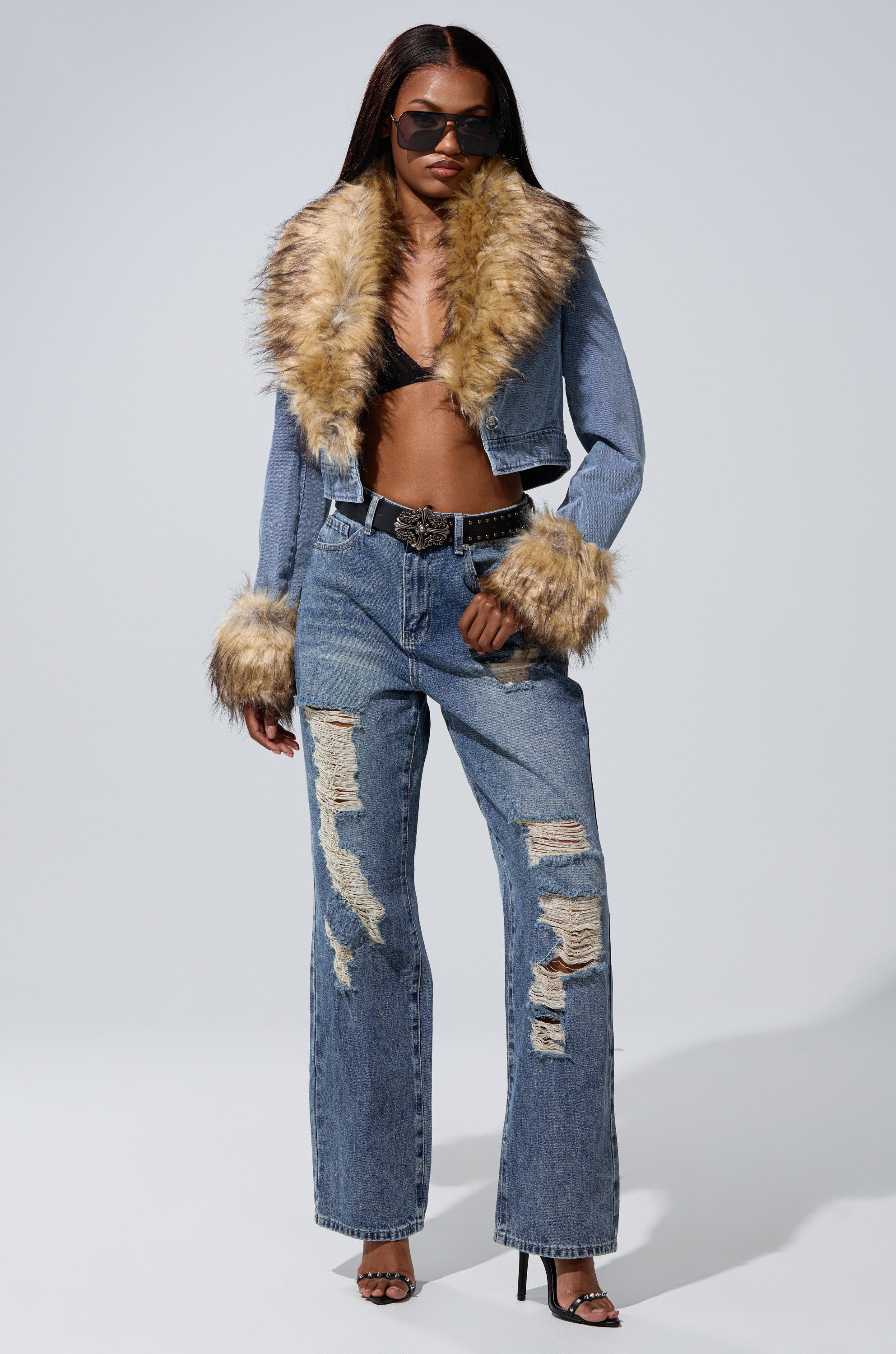 A woman poses against a plain background wearing EASILY DISTRESSED DENIM—high-rise, wide-leg, distressed jeans—paired with a cropped denim jacket with faux fur details, black bralette, heeled sandals, sunglasses, and a studded belt.