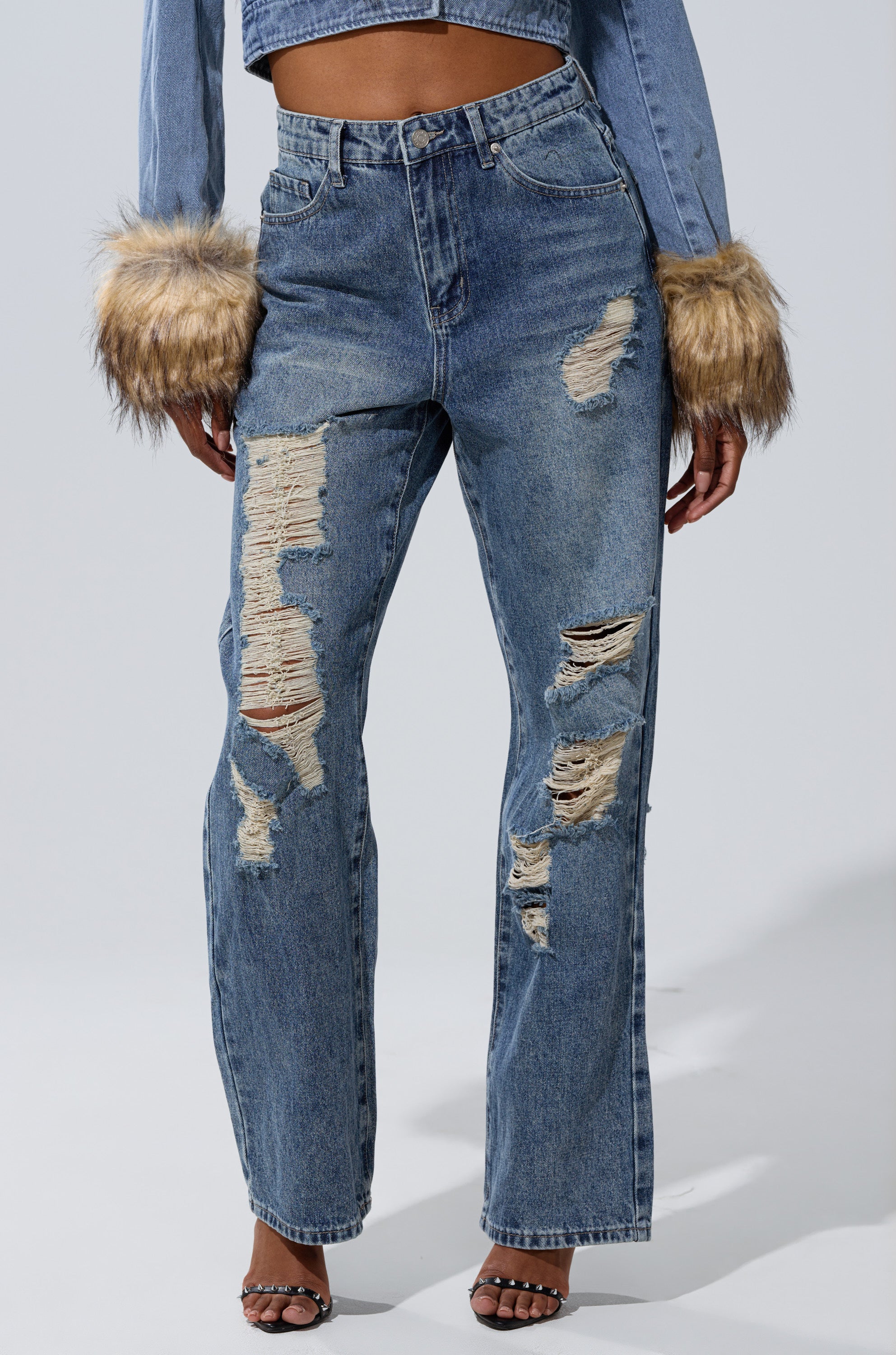 Someone wearing the EASILY DISTRESSED DENIM high-rise, blue ripped jeans with heavy distressing, styled with a cropped denim jacket with brown faux fur cuffs and open-toe heels.