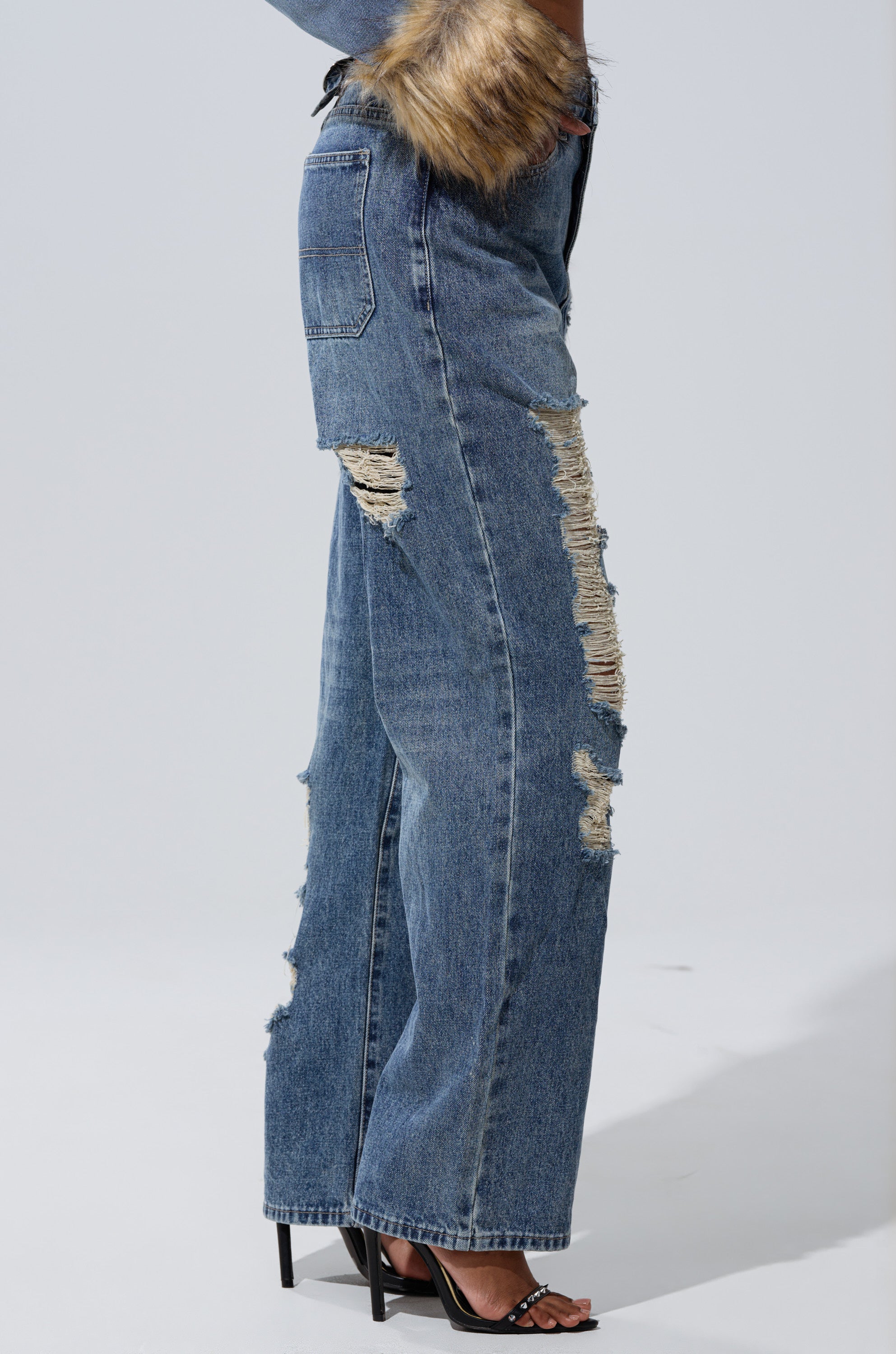 Wearing EASILY DISTRESSED DENIM high-rise jeans with multiple rips and frayed patches, black high-heeled sandals, and a jacket with faux fur cuffs, standing against a plain light background.