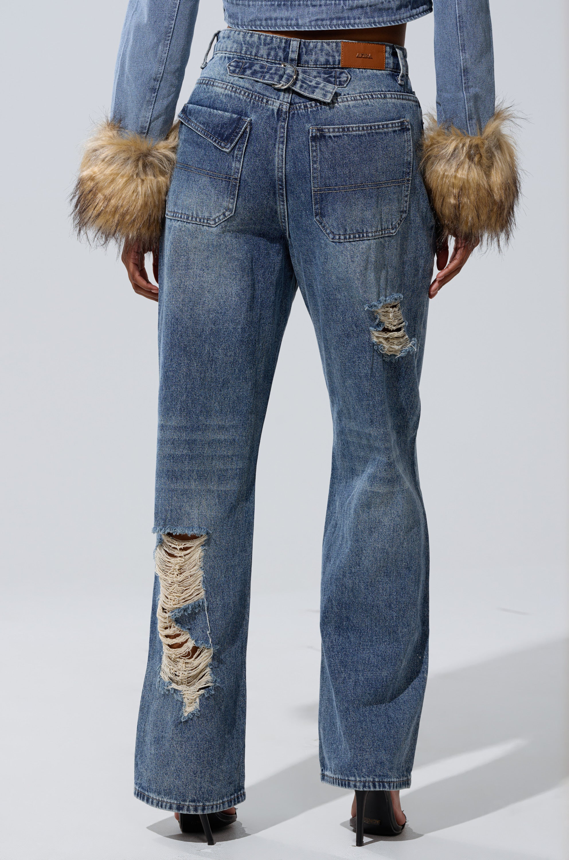 Someone is seen from behind wearing the EASILY DISTRESSED DENIM wide-leg jeans with prominent rips on one leg, paired with a denim jacket featuring faux fur cuffs.