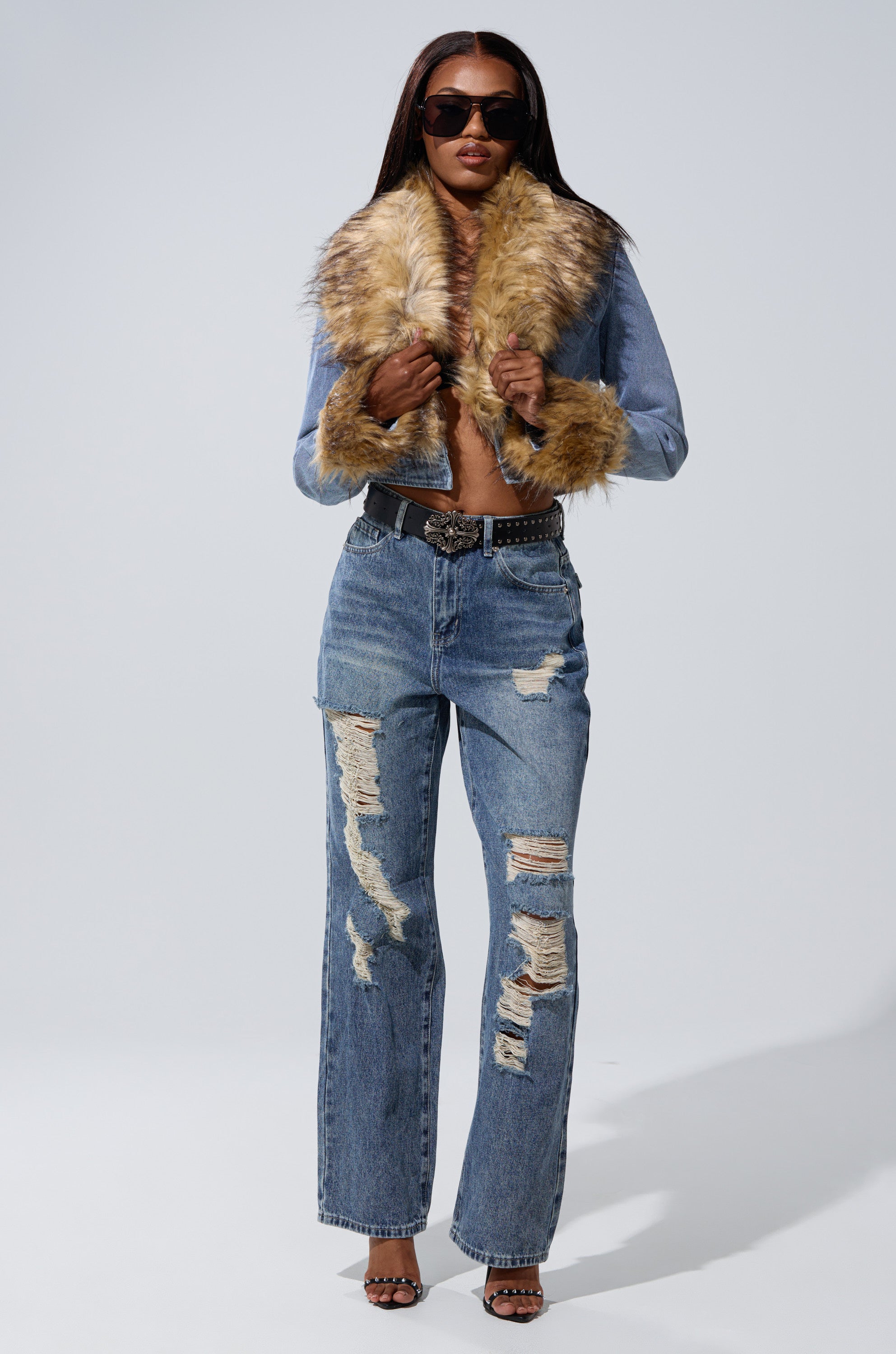 A woman poses confidently against a plain background wearing EASILY DISTRESSED DENIM jeans paired with a fur-collared denim jacket, black belt, sunglasses, and heeled sandals.