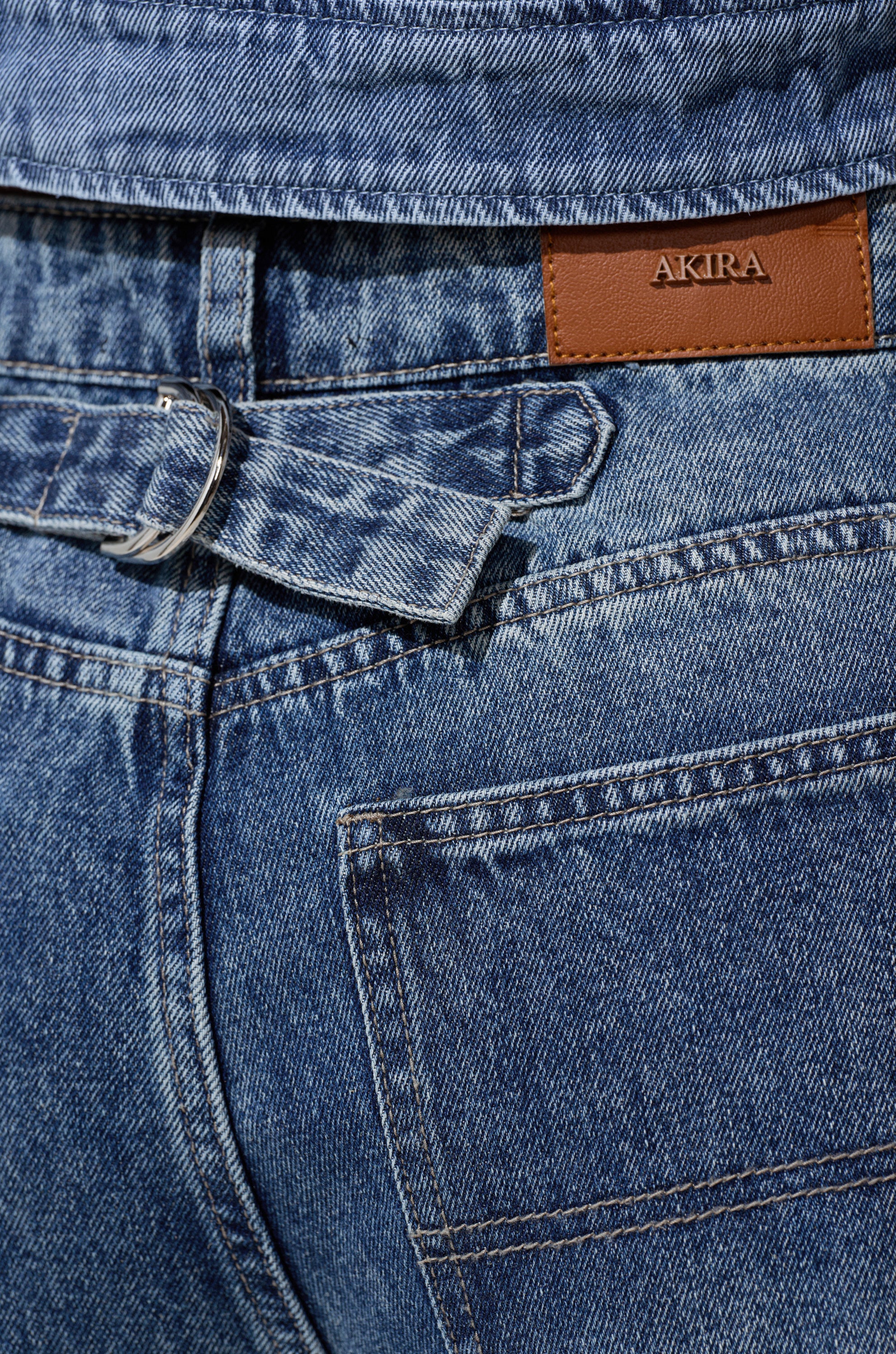 Close-up of the back of EASILY DISTRESSED DENIM jeans in blue with a brown "AKIRA" leather patch, featuring a silver-buckled belt loop and visible stitching.