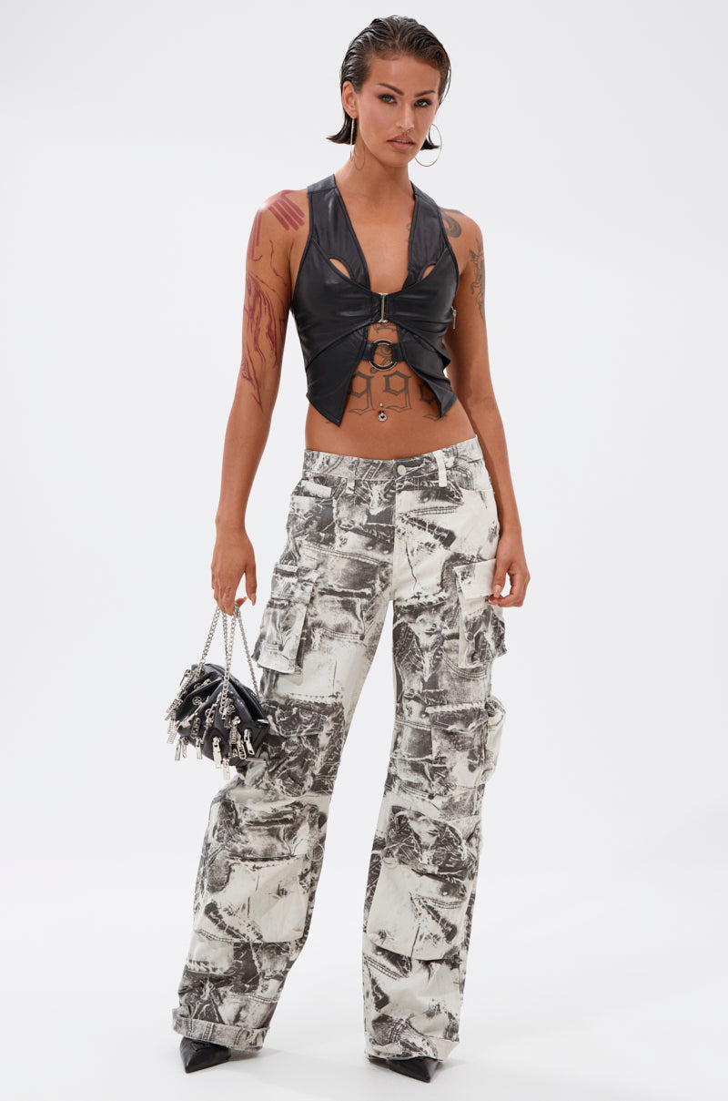 A person poses confidently in ASH OMBRE CARGO DENIM, a sleeveless black cut-out leather top, black pointed shoes, and holds a chain-strap bag. Their hair is slicked back and arm tattoos are visible. 0