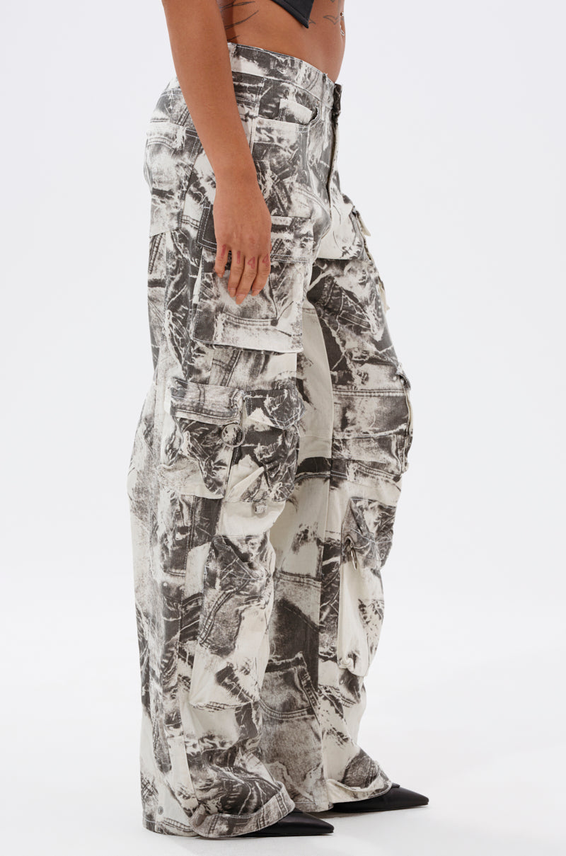 A person stands sideways wearing ASH OMBRE CARGO DENIM, featuring a high-waisted, wide-leg fit with a black and white abstract pattern, paired with black pointed-toe shoes against a plain white background. 3