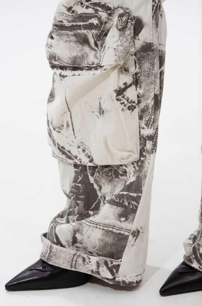 Close-up of a person wearing ASH OMBRE CARGO DENIM—loose, light-colored denim pants with large abstract black patterns, big side pockets, and pointed black shoes, set against a white background. 7