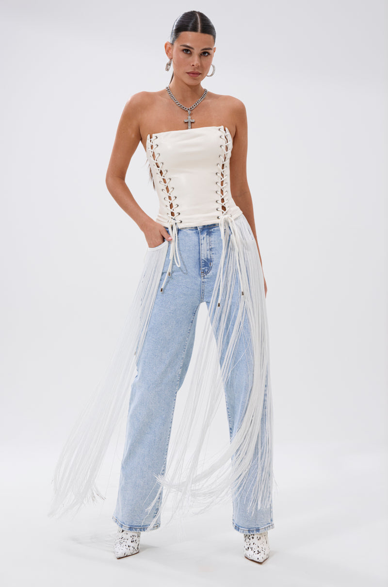 A woman poses in the FLYING AWAY FRINGE DENIM—light blue Western-style jeans with long tassel fringe—paired with a strapless white lace-up corset, white heeled boots, hoop earrings, and layered necklaces against a plain background.