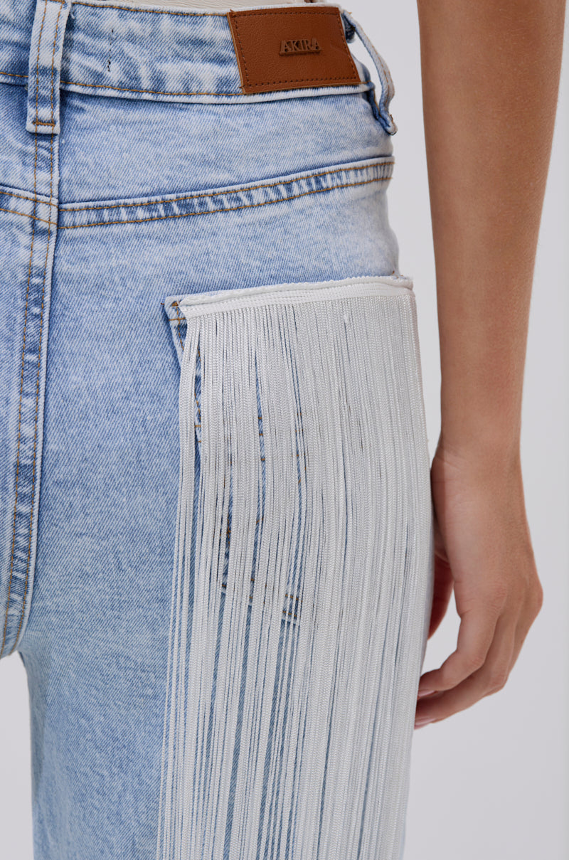 Close-up of FLYING AWAY FRINGE DENIM: light blue Western-style jeans featuring a brown leather waistband patch and long white tassel fringe detailing on the side pocket; a person’s arm appears beside the fringe.