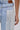 Close-up of FLYING AWAY FRINGE DENIM: light blue Western-style jeans featuring a brown leather waistband patch and long white tassel fringe detailing on the side pocket; a person’s arm appears beside the fringe.