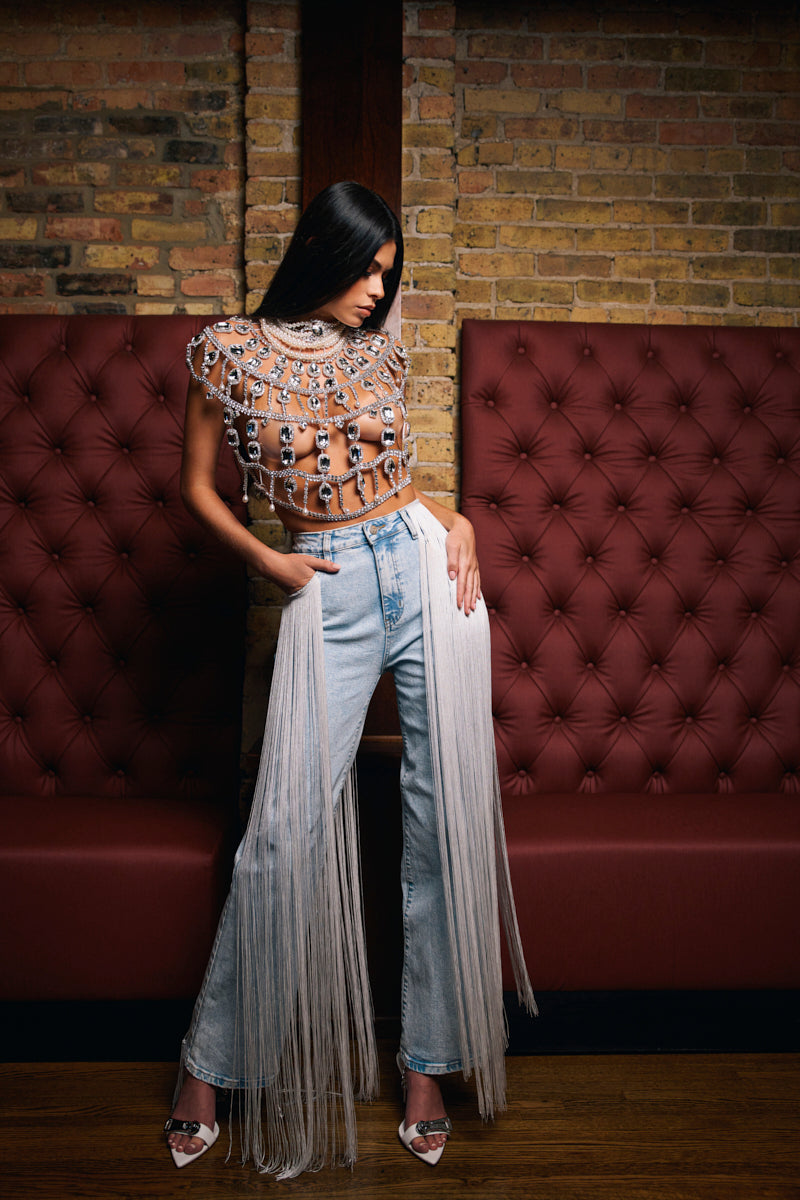 A woman poses indoors in FLYING AWAY FRINGE DENIM jeans, paired with a sheer jeweled top and white heels, standing against a brick wall with red cushioned seating and one hand on her hip as she looks down.
