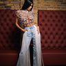 A woman poses indoors in FLYING AWAY FRINGE DENIM jeans, paired with a sheer jeweled top and white heels, standing against a brick wall with red cushioned seating and one hand on her hip as she looks down.