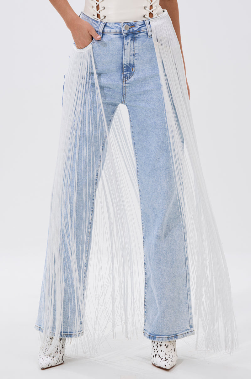Model wears FLYING AWAY FRINGE DENIM—light blue high-rise jeans with long white fringe along the sides—paired with a white top and white patterned boots against a plain white background.