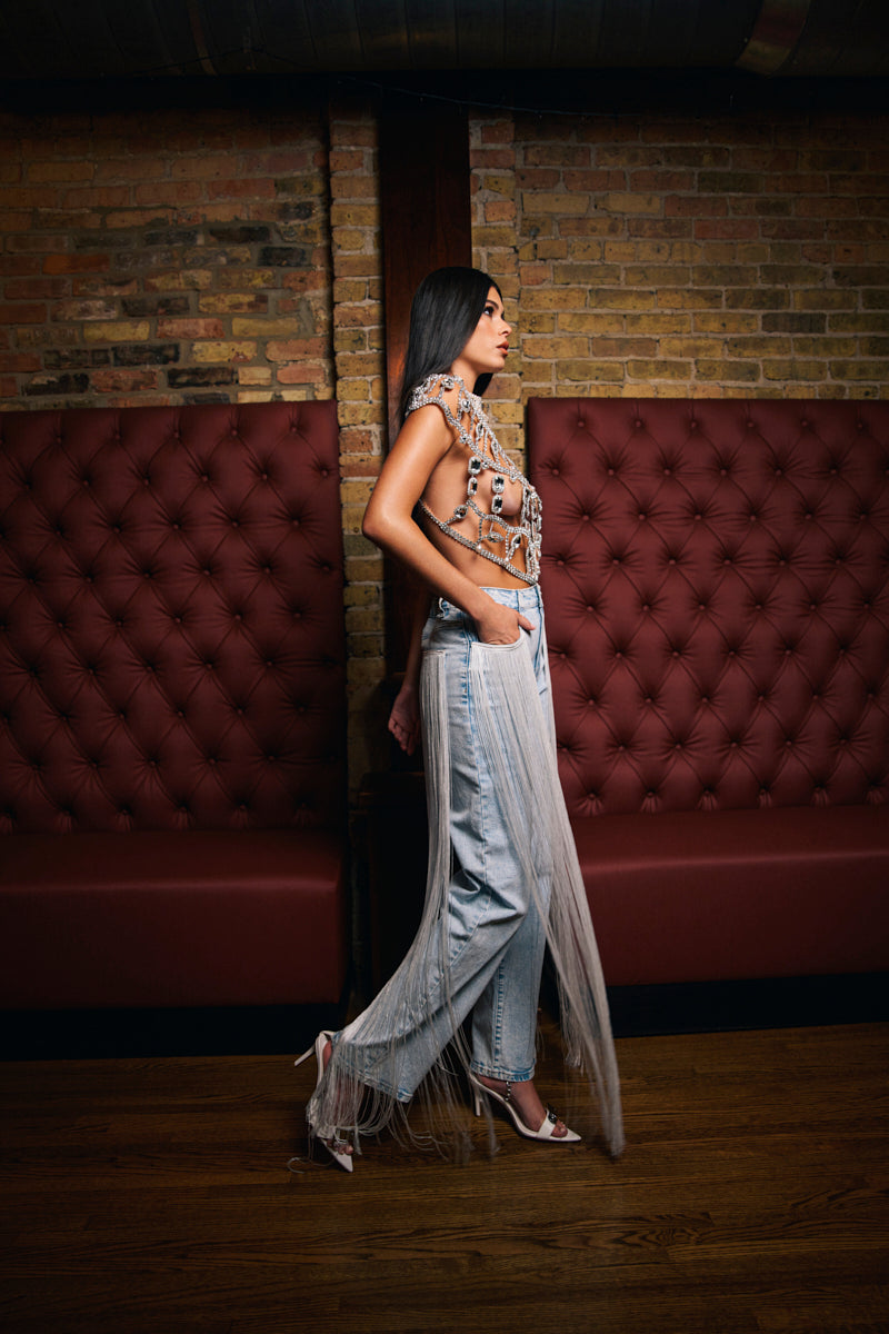A woman with long dark hair walks indoors against a brick wall with red tufted booth seating, wearing the FLYING AWAY FRINGE DENIM—high-rise, loose Western-style jeans featuring long, sheer fringe details and paired with a beaded top.