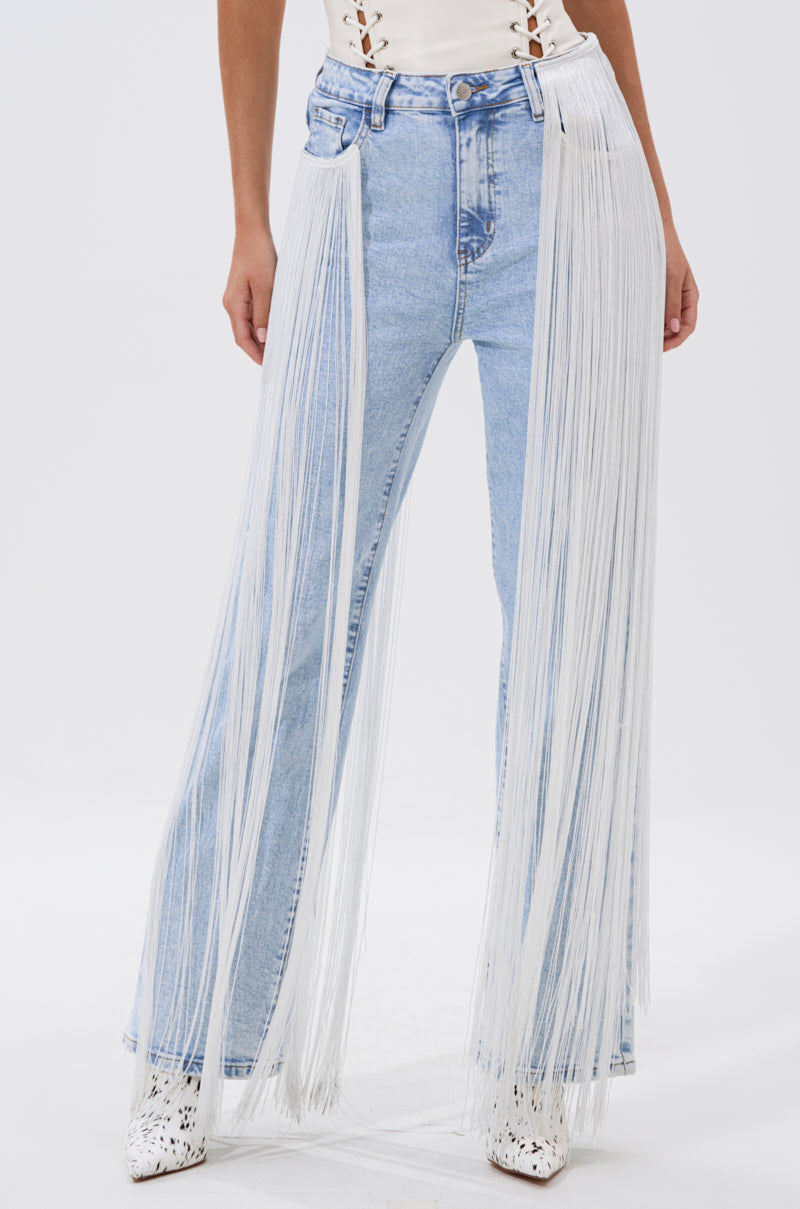 Someone wears the FLYING AWAY FRINGE DENIM—light blue, high-rise jeans with dramatic long white fringe from waist to floor—styled with a cream top and white pointed-toe shoes against a plain white background.