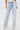 Someone wears the FLYING AWAY FRINGE DENIM—light blue, high-rise jeans with dramatic long white fringe from waist to floor—styled with a cream top and white pointed-toe shoes against a plain white background.