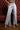 A person stands indoors in light blue FLYING AWAY FRINGE DENIM high-rise jeans and white heeled sandals, with a partially visible top. The setting has red tufted seating and a wooden floor.