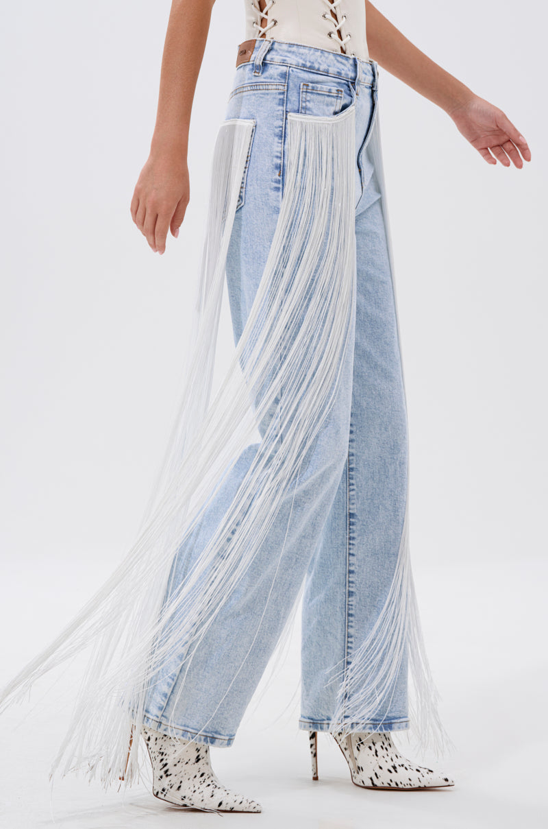 Wearing the FLYING AWAY FRINGE DENIM—light-wash, high-rise jeans with long white fringe detailing—a person pairs them with a white top and black-spotted white ankle boots, standing against a plain white background.