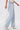 Wearing the FLYING AWAY FRINGE DENIM—light-wash, high-rise jeans with long white fringe detailing—a person pairs them with a white top and black-spotted white ankle boots, standing against a plain white background.