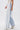 A person wears the FLYING AWAY FRINGE DENIM jeans with long white fringe, a white lace-up corset top, and spotted white high-heeled boots, standing against a plain white background.