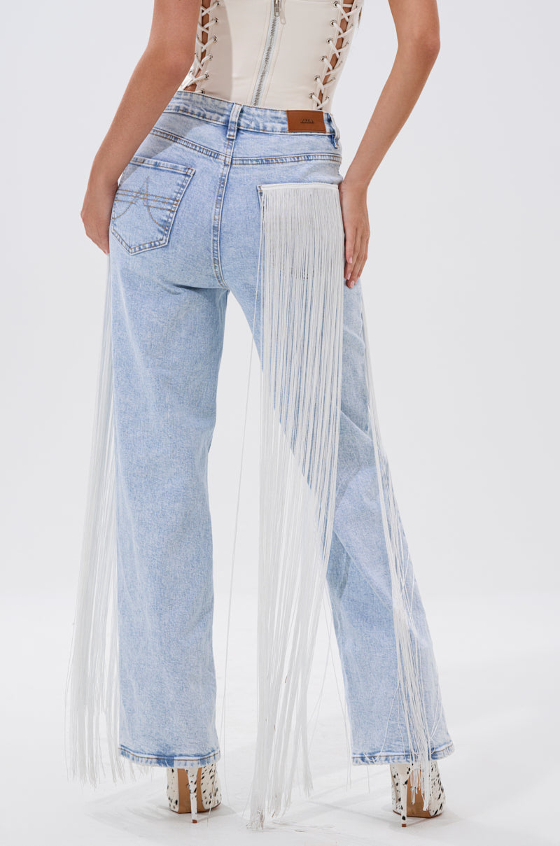 A person stands against a plain white background wearing the FLYING AWAY FRINGE DENIM—light blue, Western-style jeans with long white fringe on the sides—paired with a beige lace-up top and white heeled shoes.