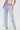 A person stands against a plain white background wearing the FLYING AWAY FRINGE DENIM—light blue, Western-style jeans with long white fringe on the sides—paired with a beige lace-up top and white heeled shoes.