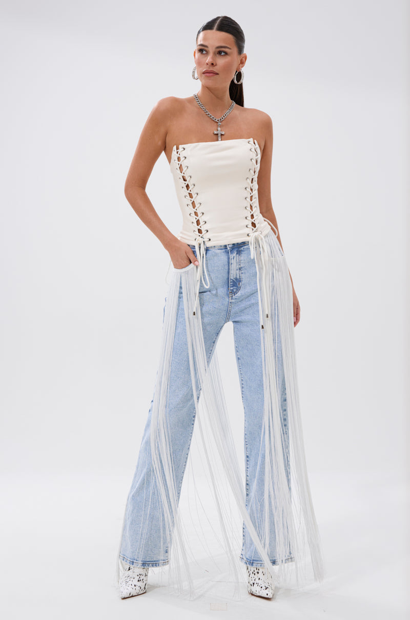 A woman stands confidently in the FLYING AWAY FRINGE DENIM—light blue high-rise jeans with white fringe—paired with a strapless white lace-up corset top, layered necklaces, and white platform shoes against a plain background.
